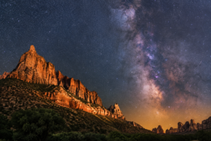 Brilliant Milky Way stretching across the night sky above towering red sandstone cliffs and rugged canyon formations, creating a breathtaking stargazing scene in the American Southwest.