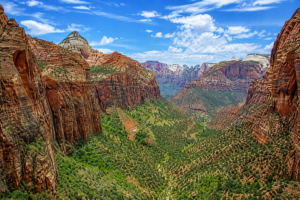 Panoramic view of towering red sandstone cliffs surrounding a lush green canyon valley beneath a bright blue sky with scattered white clouds in the American Southwest.