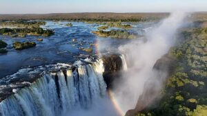 Victoria Falls Safari, Hiking, and Drone Views 1 Victoria Falls cascading into a lush rainforest, with a vibrant rainbow arching over the misty waterfall.
