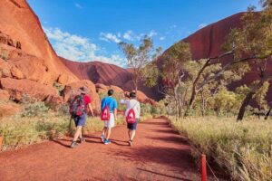 Three travelers with backpacks walking along a desert path beside towering rock wall formations at Uluru (Ayers Rock), surrounded by Australia’s red Outback landscape.