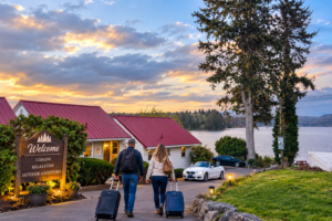 Couple walking with luggage toward lakeside lodging surrounded by evergreen trees and peaceful Oregon scenery.