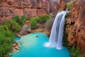 Havasu Falls Camping & Hiking Vacation 1 Spectacular waterfall cascading into a brilliant turquoise pool surrounded by towering red canyon cliffs and lush green trees in Havasu Canyon, Arizona.