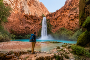 Havasu Falls Camping & Hiking Vacation 4 Hiker standing beside a turquoise pool facing a spectacular waterfall surrounded by towering red canyon cliffs at Havasu Falls in Arizona.
