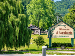 Museum of Appalachia entrance sign surrounded by lush green trees, mountain scenery, landscaped grounds, and a historic log cabin.