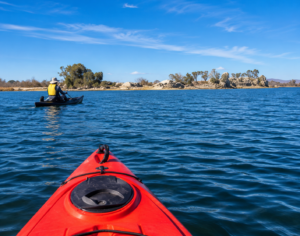 Havasu Falls Camping & Hiking Vacation 5 First-person view from a red kayak paddling across a calm blue lake toward a scenic shoreline, with another kayaker enjoying the peaceful waters under a clear blue sky.