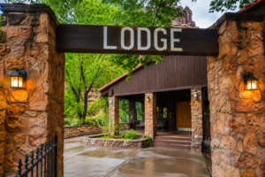 Havasu Falls Camping & Hiking Vacation 9 Rustic lodge entrance with stone pillars, warm lanterns, and a welcoming wooden sign surrounded by trees in a scenic canyon setting.