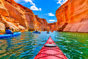 Colorful canyon kayaking adventure featuring bright red and blue kayaks paddling through crystal-clear green water surrounded by towering orange sandstone canyon walls beneath a vivid blue sky.