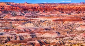 Colorful desert badlands featuring layered red, pink, purple, and cream rock formations beneath a bright blue sky.
