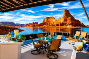 Luxury Lake Powell houseboat patio featuring outdoor dining tables, relaxing lounge chairs, colorful drinks, scenic blue water, towering red rock canyon cliffs, and breathtaking Southwest lake views beneath a vibrant sky.
