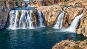 Wide view of multiple waterfalls cascading over rugged canyon cliffs into a deep blue-green pool, with a hiker observing the dramatic landscape.