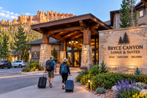 Bryce Canyon Utah Vacation 6 Travelers carrying backpacks and rolling luggage walk toward a welcoming lodge entrance surrounded by pine trees and Bryce Canyon’s red-orange rock formations in Utah.