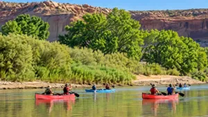 Lower Calf Creek Utah Vacation 5 Canoers and kayakers paddle along a calm river surrounded by green vegetation and towering sandstone canyon walls.