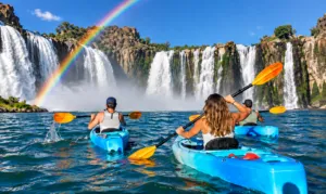 Three kayakers paddling across blue water toward massive cascading waterfalls beneath a colorful rainbow and bright blue sky.