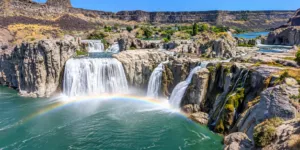 Wide panoramic view of Shoshone Falls cascading over rugged canyon cliffs into blue-green water with a colorful rainbow rising through the mist.
