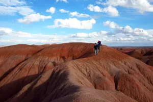 Hikers walking across colorful red badland formations in the Painted Desert of Arizona beneath a bright blue sky.