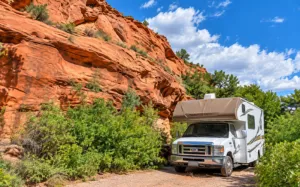 Lower Calf Creek Utah Vacation 6 White RV parked beneath towering red sandstone cliffs surrounded by green desert vegetation under a bright blue sky.