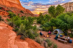 Lower Calf Creek Utah Vacation 4 Scenic canyon campsite with a tent, campfire, picnic table, and camping gear surrounded by red-rock cliffs and lush green vegetation.