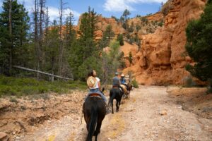 Bryce Canyon Utah Vacation 5 people horseback riding on a path near trees and large canyon walls and a partly cloudy day.