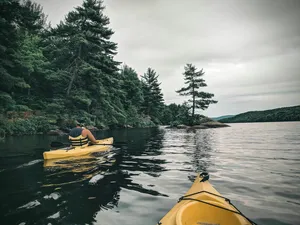 Kayaker wearing a yellow life jacket paddling a yellow kayak across calm lake waters, surrounded by dense evergreen forests, rocky shorelines, small tree-covered islands, and distant wooded hills beneath a cloudy sky.