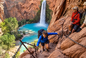 Hiker using safety chains on a steep canyon trail overlooking turquoise waterfalls and pools at Havasu Falls, Arizona.