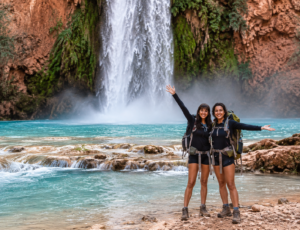 Two hikers standing beside turquoise water with a powerful waterfall cascading over red-rock cliffs at Havasu Falls, Arizona.