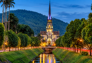 Illuminated Gothic cathedral in Petrópolis, Brazil, framed by lush trees and mountains with evening lights reflecting in the water.