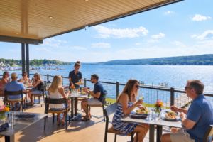 Explore New York’s Scenic Finger Lakes Region 5 Visitors enjoying a meal on a sunny waterfront restaurant patio overlooking a scenic Finger Lakes lake in New York.