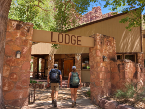 Two hikers with backpacks walking toward a rustic lodge entrance surrounded by red-rock canyon scenery near Havasu Falls, Arizona.