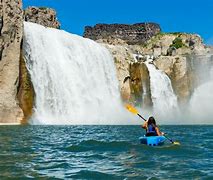 Person canoeing at the bottom of a large waterfall with steep cliffs on a blue sky day.