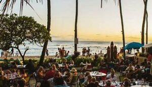 people eating at a restaurant at tables sitting in chairs near the ocean