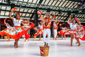 Colorful cultural dance performance featuring dancers in traditional-style costumes performing with vibrant skirts, hats, and handcrafted props.