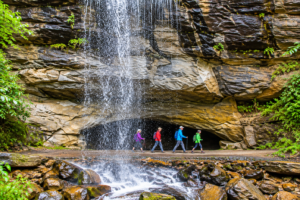 Bridal Veil Falls Utah: Hiking, Waterfalls & Outdoor Adventures 3 Hikers walking behind a cascading waterfall along a rocky trail surrounded by lush greenery.
