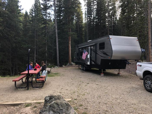 Family enjoying an RV camping adventure at South Fork Campground in Bighorn National Forest, Wyoming, with a large travel trailer, picnic table, and towering evergreen trees.