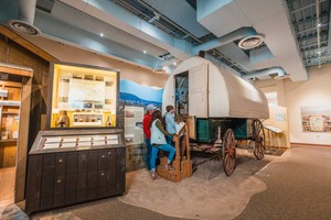 Visitors exploring a Wyoming history museum exhibit featuring a historic covered wagon, informational displays, and artifacts showcasing pioneer life and Western heritage.