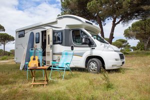 White motorhome parked at a peaceful grassy campsite surrounded by trees, with turquoise camping chairs, a small wooden table, guitar, and outdoor recreational gear beside the RV.