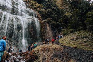 Group of hikers exploring a scenic waterfall surrounded by steep rocky cliffs and dense forest, with cascading water flowing down the rock face and visitors walking along a rugged trail.