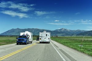 RV motorhome and pickup truck towing a camper along a scenic highway surrounded by green fields, distant mountain peaks, and blue skies.