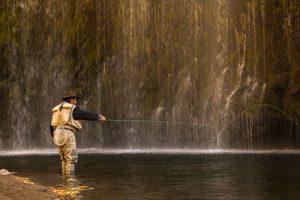 Fly fisherman wearing chest waders and a brown hat casting a fishing line into a shallow pool beneath a beautiful cascading waterfall surrounded by rocky cliffs.