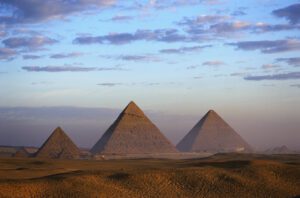 Three large pyramids of Egypt with three smaller pyramids nearby, surrounded by desert sands under a partly cloudy sky.