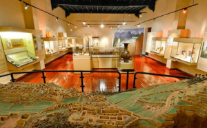 Museum interior in Cusco, Peru, featuring illuminated display cases with archaeological artifacts and a detailed scale model of an ancient Andean settlement.
