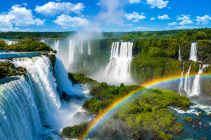 Iguazu Falls Photography Tours 1 Panoramic view of Iguazu Falls with multiple cascading waterfalls, lush rainforest, rising mist, and a vibrant rainbow stretching across the river.