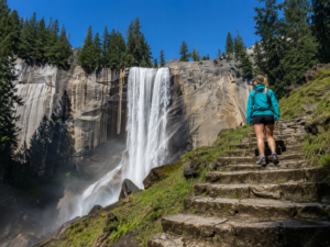 Hiker climbing stone steps toward Vernal Fall in Yosemite National Park, California, with a powerful waterfall cascading over granite cliffs surrounded by evergreen forest.