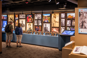 Two visitors wearing backpacks exploring a museum history exhibit featuring vintage photographs, historical portraits, interactive touchscreen displays, and a large “What’s Your Story?” sign.