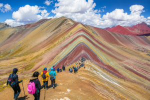 Hikers trekking along the colorful striped slopes of Rainbow Mountain near Cusco, Peru, beneath a bright blue sky.