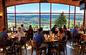 Guests enjoying meals at wooden tables inside a rustic restaurant with exposed brick walls, large panoramic windows, and breathtaking views of a scenic river canyon surrounded by green hills and rocky cliffs.