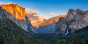 Wide panoramic view of Yosemite Valley in Yosemite National Park, California, featuring towering granite cliffs, a majestic waterfall, dense evergreen forests, and distant mountain peaks glowing in the warm light of sunrise.