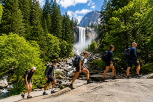 Group of hikers crossing granite rocks toward Vernal Fall in Yosemite National Park, California, with a towering waterfall, evergreen forest, and mountain scenery.