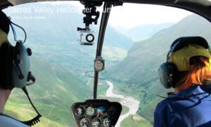 Aerial view of the Sacred Valley near Cusco, Peru, taken from inside a helicopter cockpit, showing dramatic cliffs, winding river, and expansive valley scenery below.
