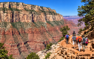 Group of hikers walking along a rocky canyon trail surrounded by towering red and tan cliffs, rugged desert terrain, scattered evergreen trees, and expansive canyon views.