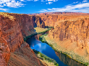 Clear panoramic view of a winding river flowing through towering red-orange canyon walls beneath a bright blue sky with scattered clouds.